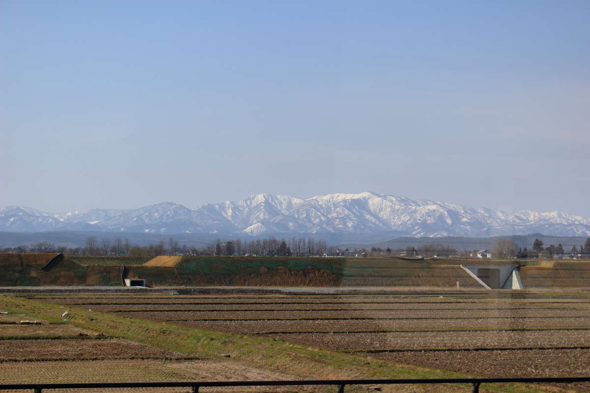 田園風景の向こうに雪をかぶった山並みを望む「フラワー長井線」の車窓